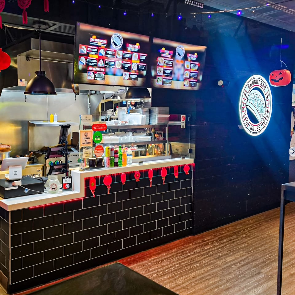 Interior of Chubby Bao House, featuring a counter with menu screens, kitchen, and a neon logo.