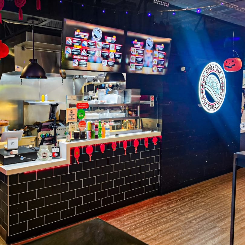 Interior of Chubby Bao House, featuring a counter with menu screens, kitchen, and a neon logo.