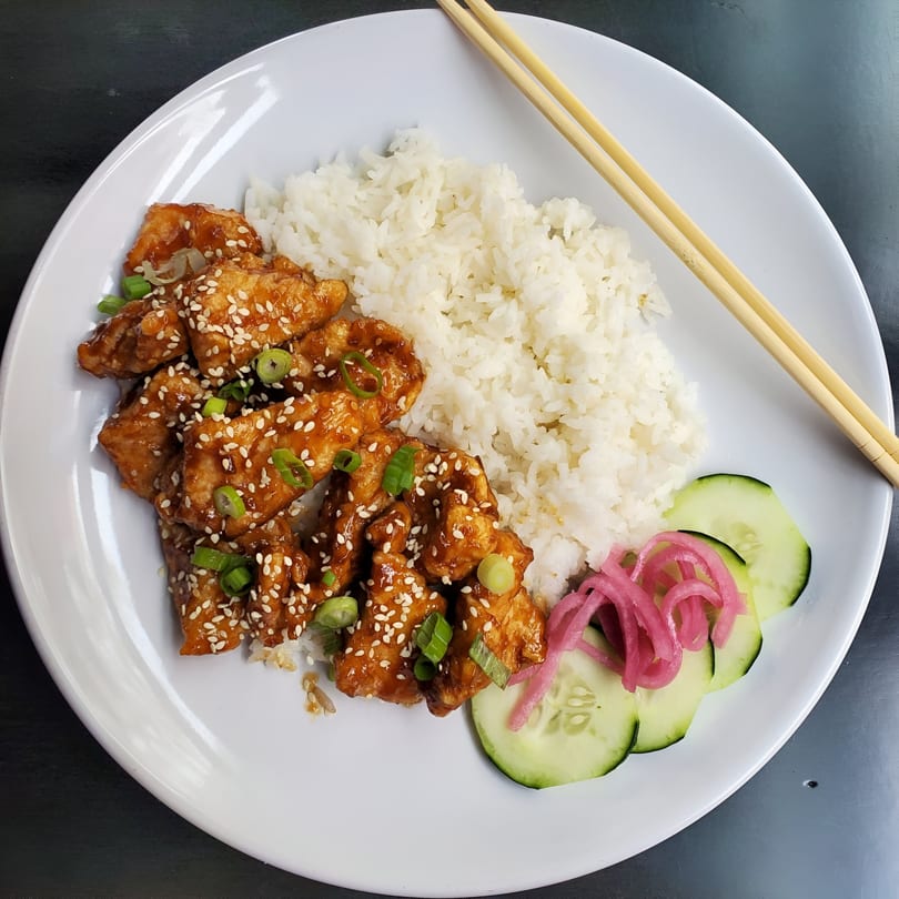 Chubby Bao House's sesame chicken with rice, pickled onions, and cucumber slices, with chopsticks.